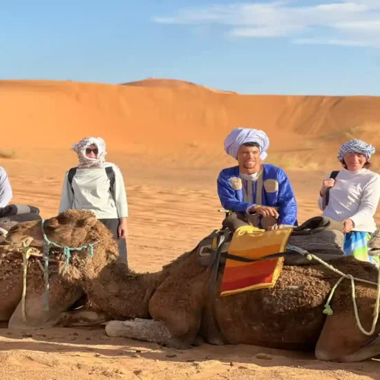 Travelers wearing traditional headscarves posing with camels and a local guide in the golden sand dunes of Merzouga Sahara Desert. 3 days tour from Fes to Marrakech