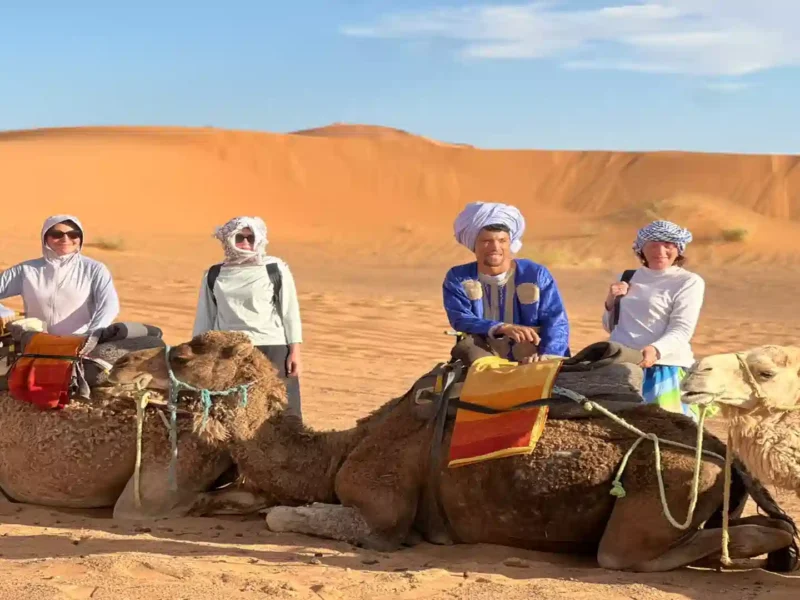 Travelers wearing traditional headscarves posing with camels and a local guide in the golden sand dunes of Merzouga Sahara Desert. 3 days tour from Fes to Marrakech