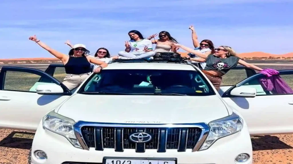 Group of friends posing cheerfully around and on top of a white Toyota 4x4 tour vehicle in the Moroccan desert. 3 days tour from Fes to Marrakech