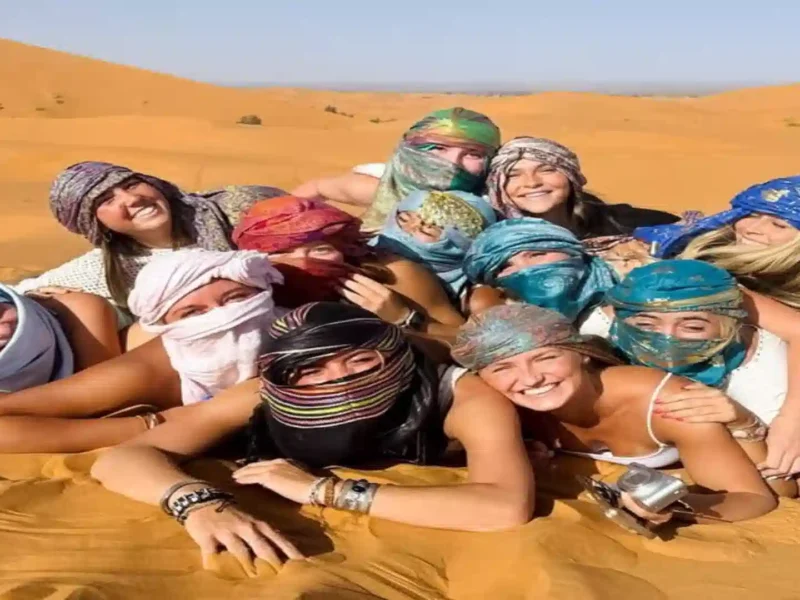 Group of happy travelers lying together on the golden sand dunes of the Merzouga Sahara Desert wearing colorful headscarves.5 days tour from Marrakech to Merzouga- Over Morocco Tours