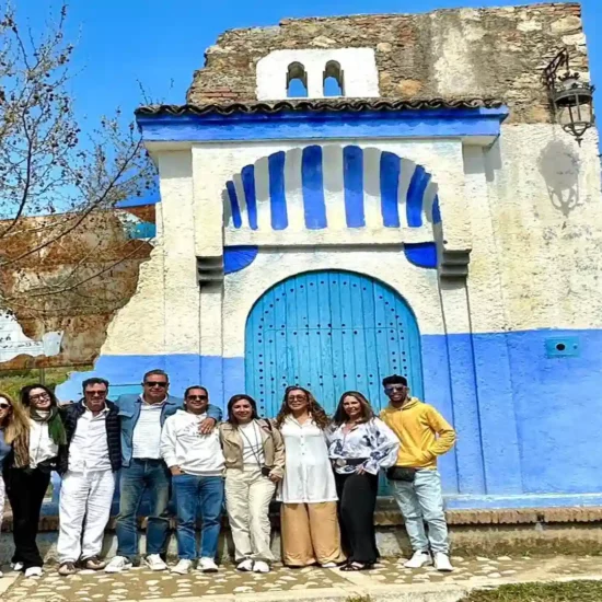 : Group of tourists posing in front of a traditional blue and white painted stone structure near Chefchaouen with green Rif Mountains in the background. 10 day Morocco tour from Marrakech