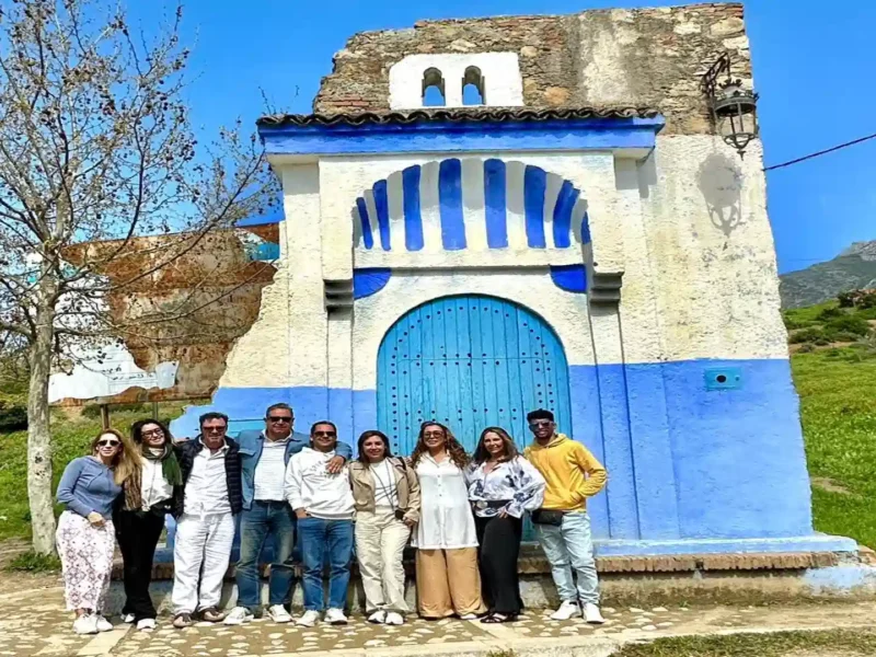 : Group of tourists posing in front of a traditional blue and white painted stone structure near Chefchaouen with green Rif Mountains in the background. 10 day Morocco tour from Marrakech