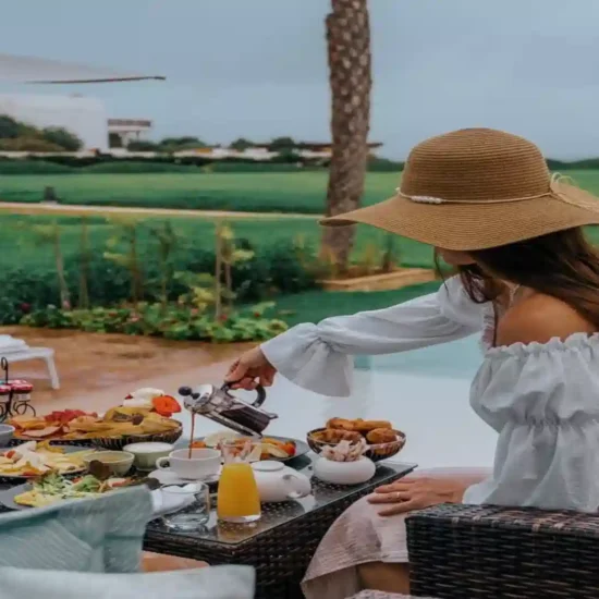 Woman in a straw hat pouring tea at a poolside breakfast table with a lush green golf course view, part of an 8 days tour From Agadir to Marrakech Essaouira tour.