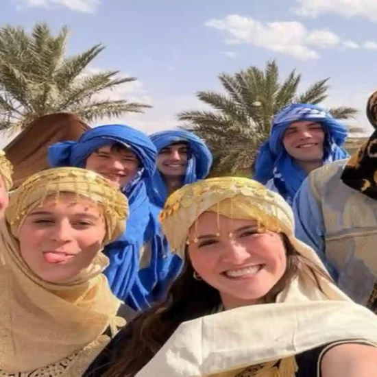Happy group of travelers wearing traditional Moroccan Amazigh (Berber) clothing and headscarves smiling for a selfie in a palm oasis.4 days tour from Marrakech to Fes