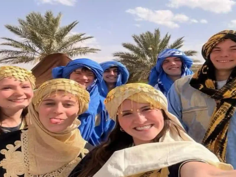 Happy group of travelers wearing traditional Moroccan Amazigh (Berber) clothing and headscarves smiling for a selfie in a palm oasis.4 days tour from Marrakech to Fes
