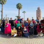 A group of tourists posing for a photo in front of the Koutoubia Mosque minaret and palm trees in Marrakech, accompanied by traditional water sellers. things to do in Marrakech