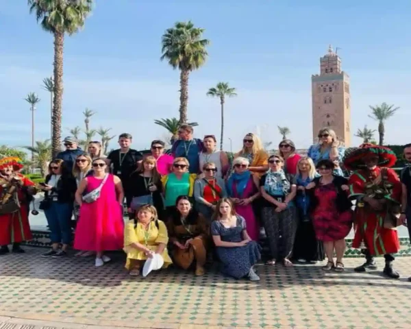A group of tourists posing for a photo in front of the Koutoubia Mosque minaret and palm trees in Marrakech, accompanied by traditional water sellers. things to do in Marrakech