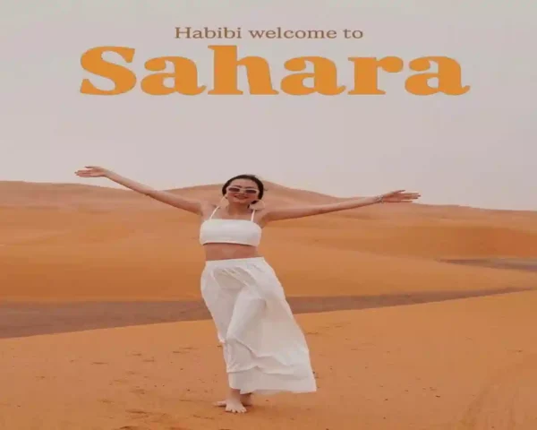 A woman in a white outfit standing on golden sand dunes with the text Habibi welcome to Sahara on a Morocco in December