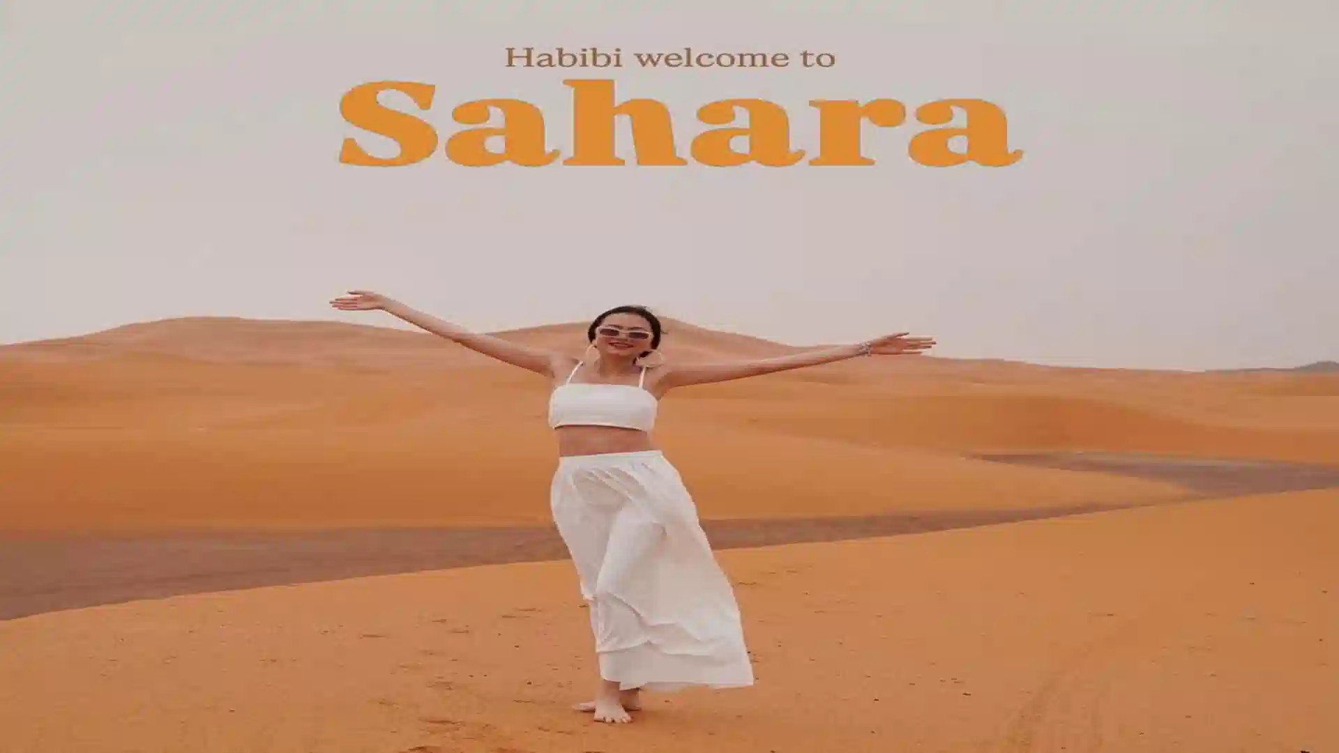 A woman in a white outfit standing on golden sand dunes with the text Habibi welcome to Sahara on a Morocco in December