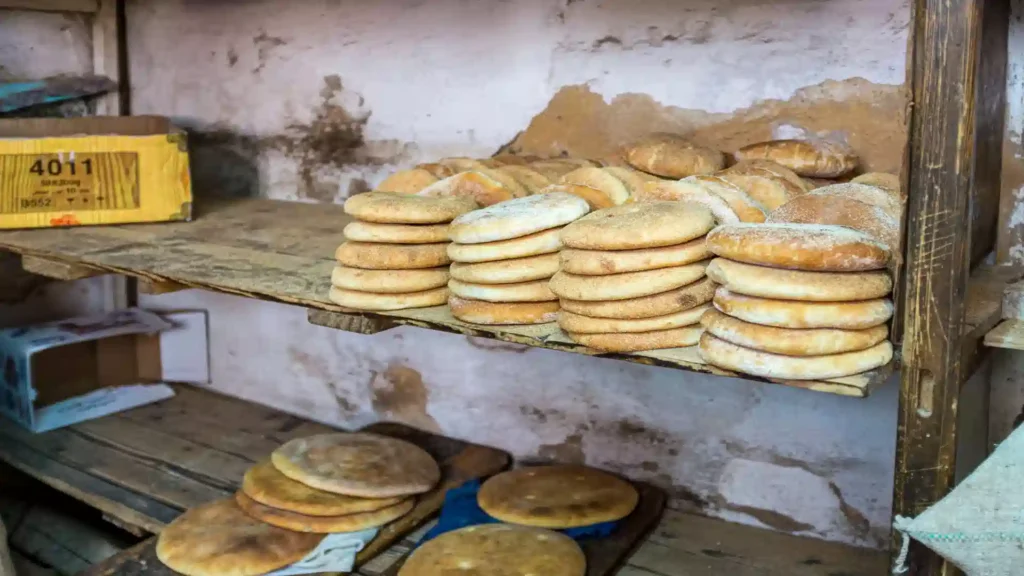 Stacks of freshly baked, round Moroccan khobz flatbread resting on rustic wooden shelves inside a traditional market bakery in Morocco.