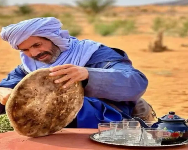 A man wearing a light blue turban and blue robe dusts sand from a large, circular Moroccan flatbread (khobz) that was baked in the Sahara desert sand.