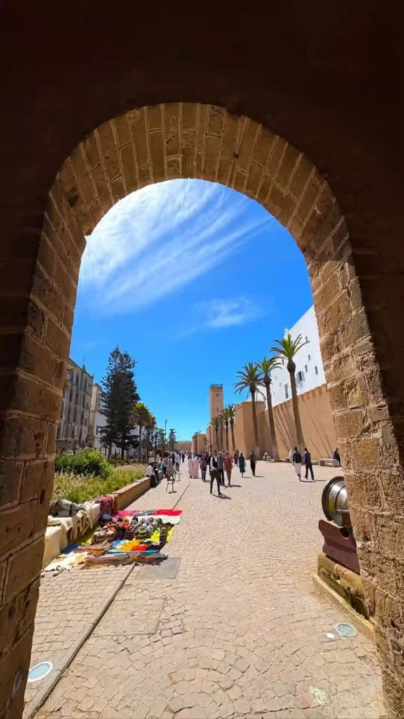 Looking through a tall stone archway toward palm trees, local vendors, and visitors walking through Essaouira.things to do in Essaouira