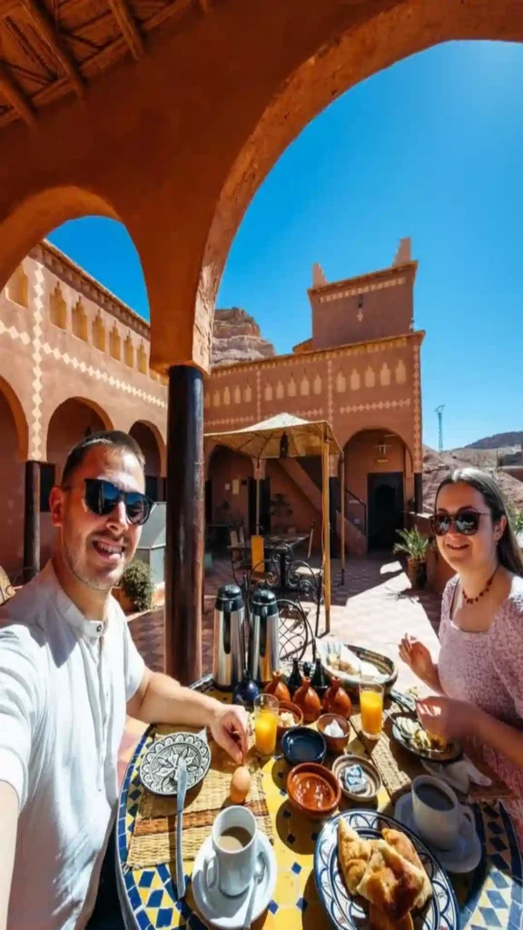 A couple enjoying a traditional Moroccan breakfast on a shaded hotel terrace with Aït Benhaddou visible in the background. where to stay in Morocco