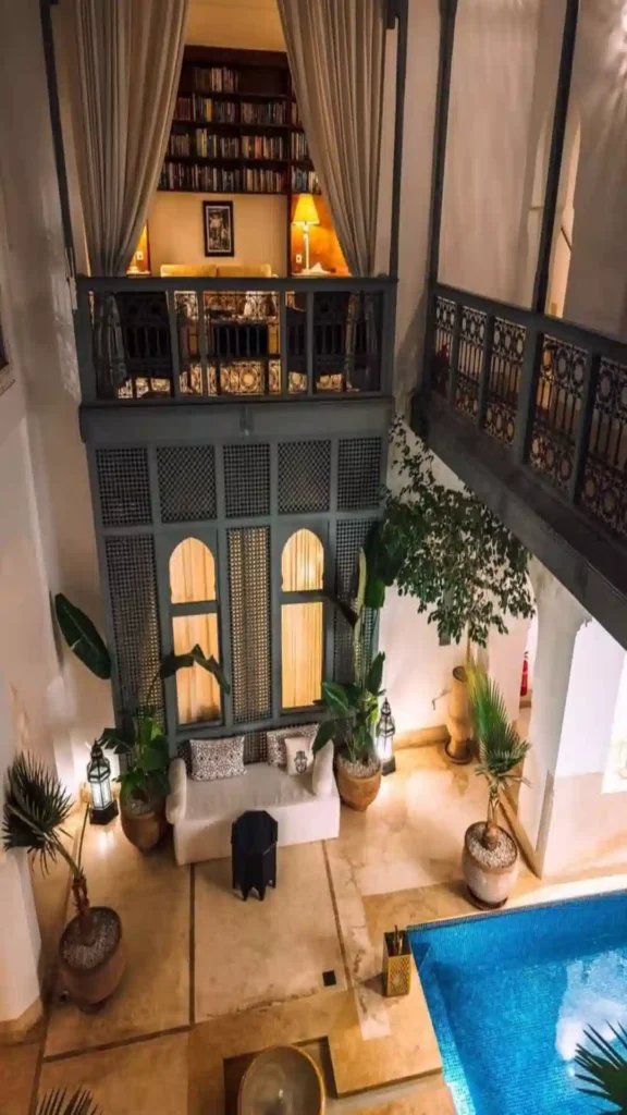 Indoor view looking down at a traditional Moroccan riad courtyard featuring a plunge pool, potted palms, and a second-floor balcony library.where to stay in Morocco.