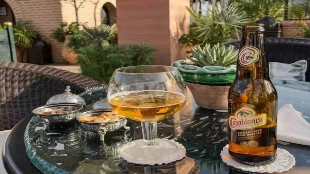 A cold bottle of Casablanca Premium Lager next to a snifter glass of beer and silver snack bowls on a glass table. can you drink alcohole in Morocco