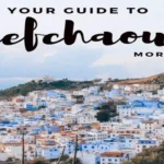 View of Chefchaouen's blue-and-white medina cascading down the hillside framed by green tree leaves in the foreground. why is Chefchaouen blue?