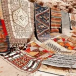 An outdoor display of colorful handmade Moroccan rug carpet and flat-woven Kilims resting against desert rocks and stone architecture.