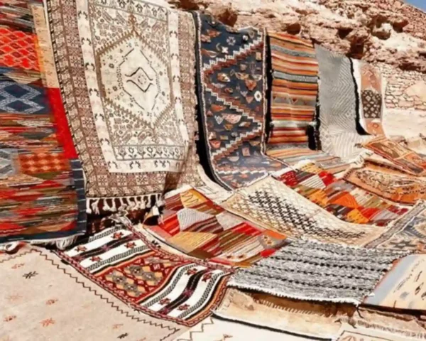 An outdoor display of colorful handmade Moroccan rug carpet and flat-woven Kilims resting against desert rocks and stone architecture.