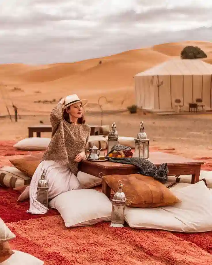 A woman sitting by a low table on plush floor cushions outside a desert tent during a Morocco luxury travel itinerary.