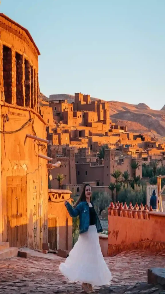 A female traveler in a denim jacket and white skirt twirling in front of the mud-brick fortifications of Ksar Aït Benhaddou.where to stay in Morocco.