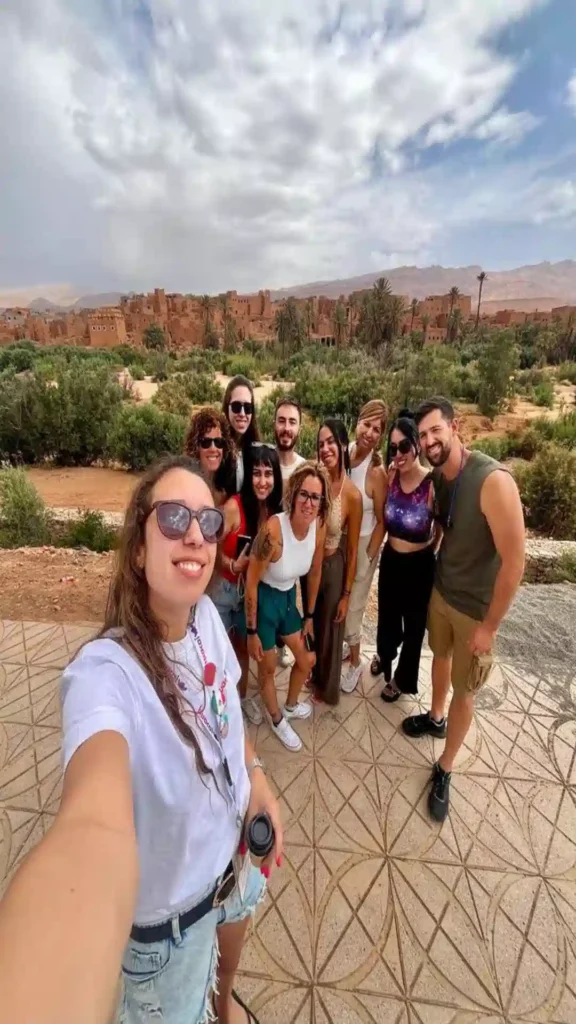 A diverse group of travelers taking a wide selfie on a tiled terrace overlooking a palm grove oasis and ancient kasbah on a Morocco desert tour from Marrakech.