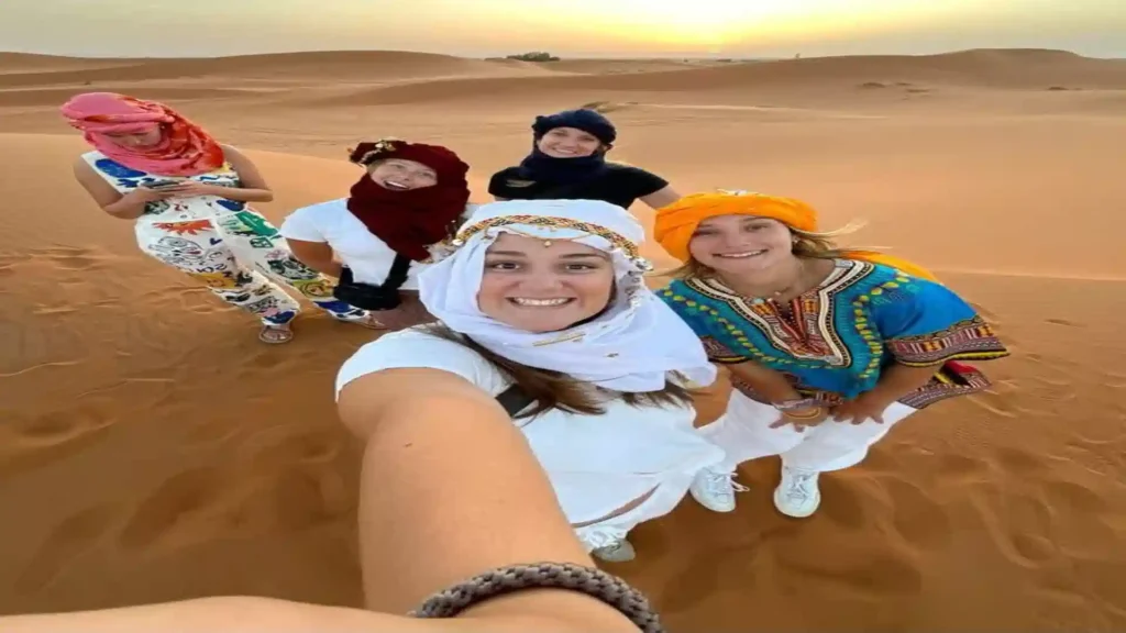 A group of five women wearing colorful headscarves posing for a wide-angle selfie on top of a sand dune in Merzouga Morocco.