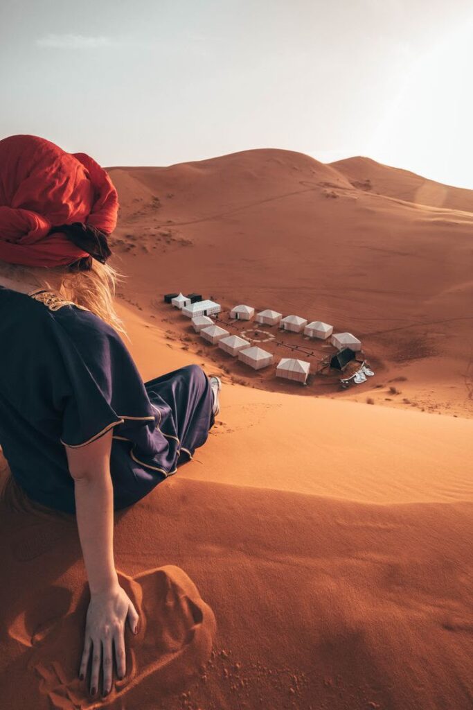 A traveler sitting atop a high sand dune overlooking a luxury desert camp below during a luxury travel Morocco excursion.