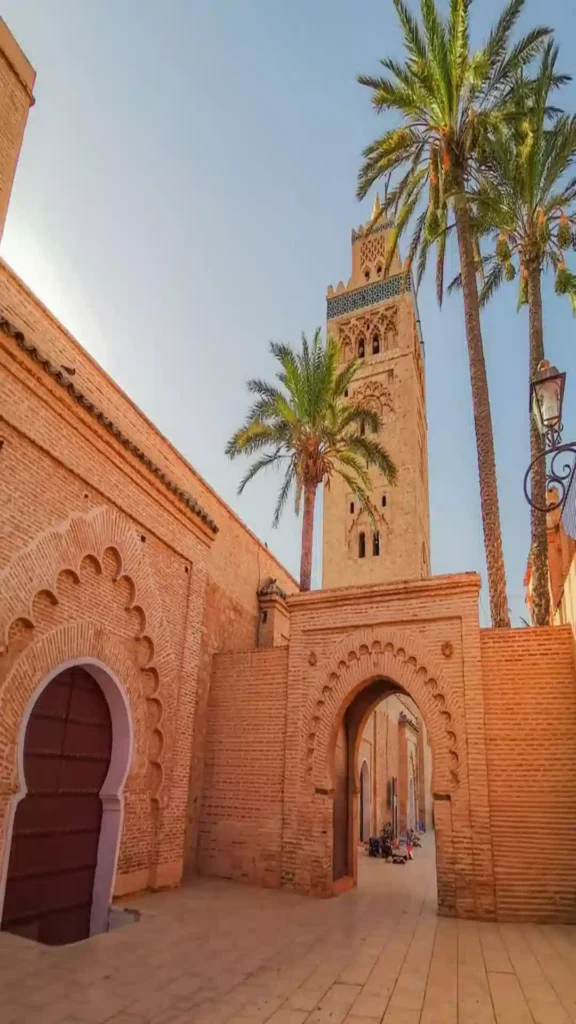 The iconic minaret of the Koutoubia Mosque framed by palm trees and terracotta arches before departing on a Morocco desert tour from Marrakech.
