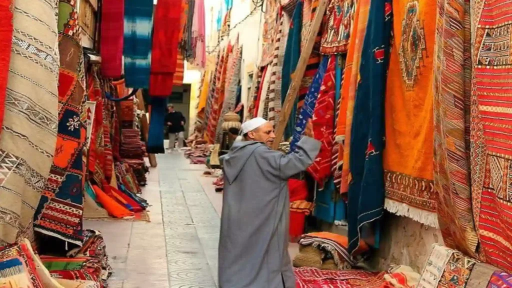 A local merchant wearing a traditional djellaba hanging vibrant woven rugs and textiles in a narrow Moroccan carpet medina souk street.