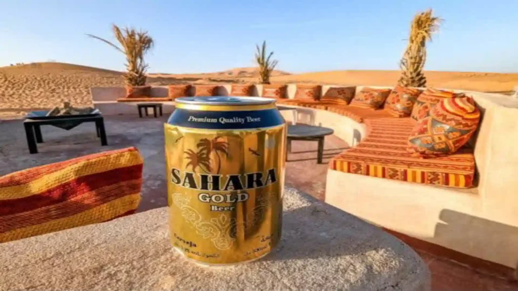 A golden can of Sahara Gold beer sitting on a stone ledge with traditional desert seating cushions and sand dunes in the background. Alcohol in Morocco