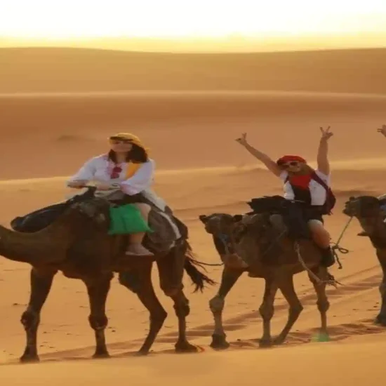 Three happy women wearing headscarves riding camels in a line across golden sand dunes during a desert sunset in Erg Chebbi, Morocco.1 week in Morocco
