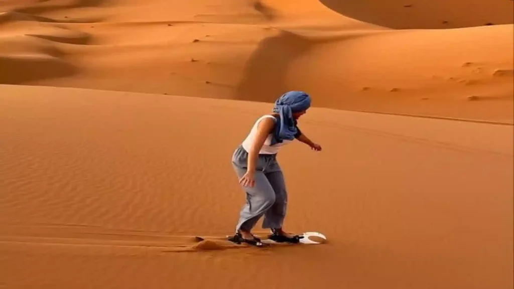A woman wearing a blue headscarf sandboarding down a steep orange dune in the Erg Chebbi Dunes.