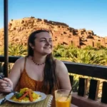 A woman dining on traditional tagine at a rooftop restaurant terrace overlooking a palm oasis and earthen kasbah village. where to stay in Morocco.