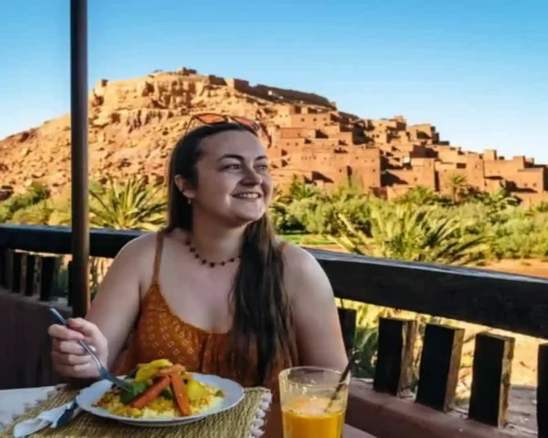 A woman dining on traditional tagine at a rooftop restaurant terrace overlooking a palm oasis and earthen kasbah village. where to stay in Morocco.