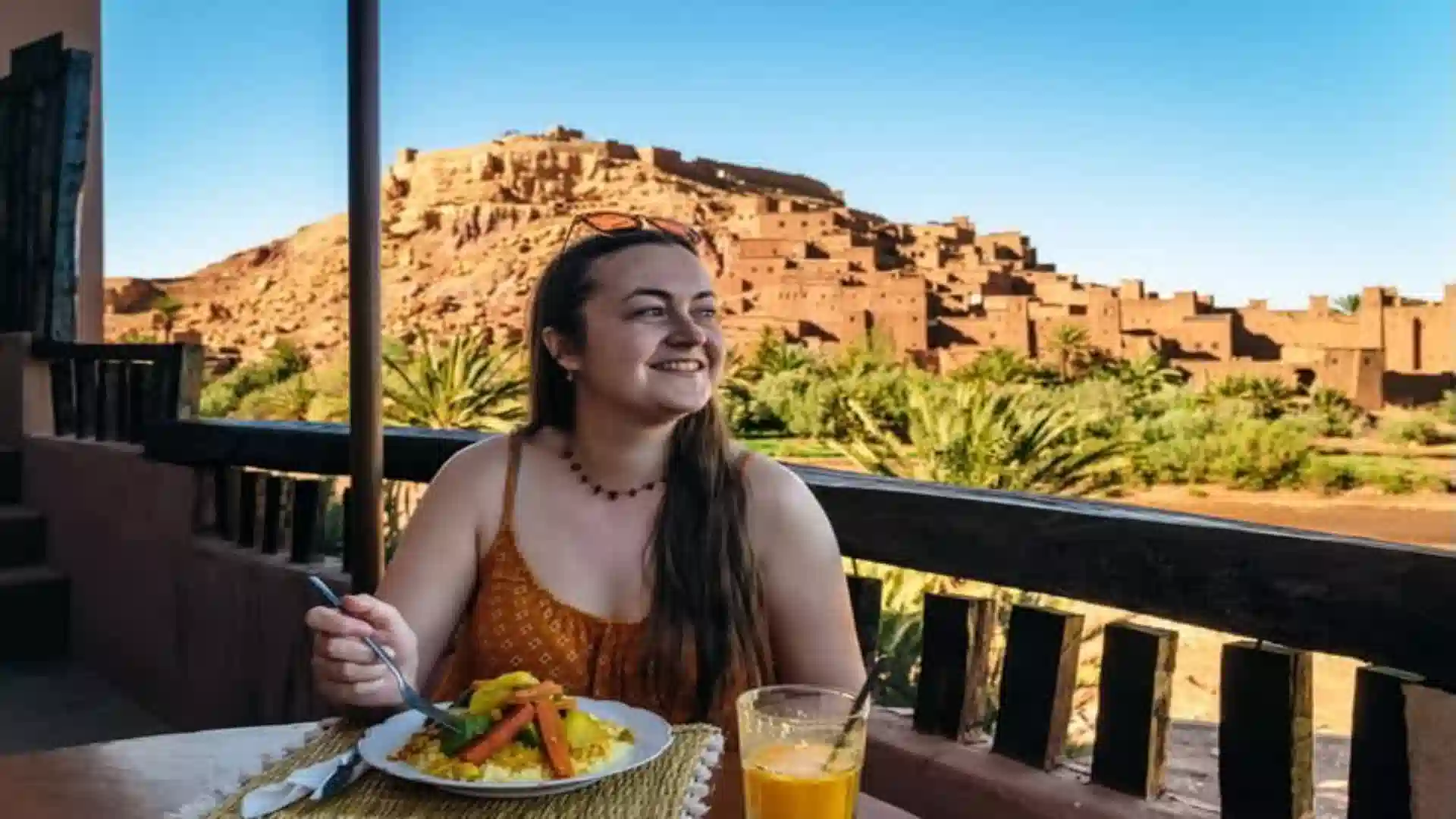 A woman dining on traditional tagine at a rooftop restaurant terrace overlooking a palm oasis and earthen kasbah village. where to stay in Morocco.