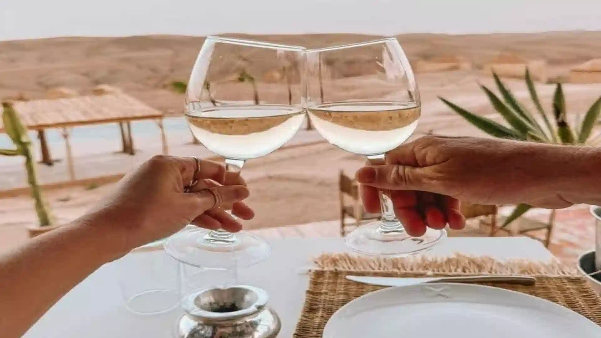 Two wine glasses clinking together with a luxury desert resort and pool in the background during luxury travel Morocco.
