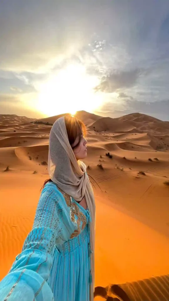 A traveler taking a selfie in a light blue dress and headscarf during sunset at Erg Chebbi sand dunes in the Sahara Desert Morocco.