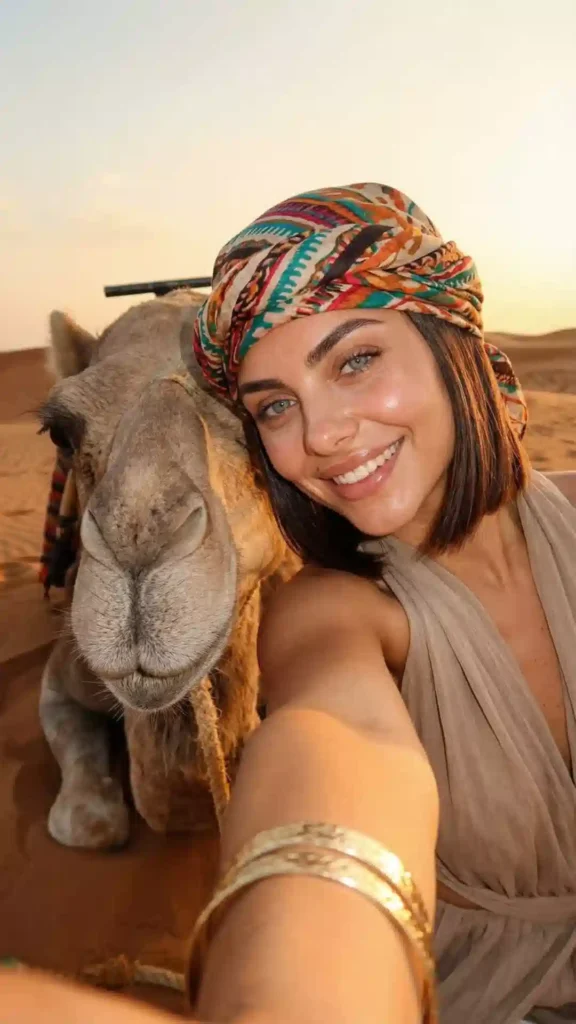 A woman in a colorful headscarf taking a selfie next to a resting camel on a camel trek in Morocco.