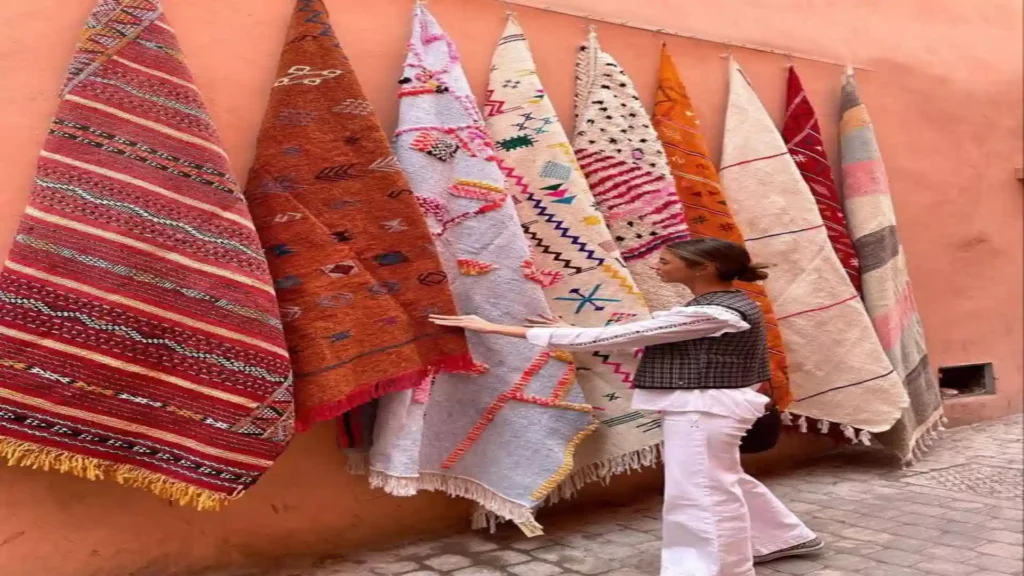 A traveler in white clothing examining colorful handwoven Moroccan carpet Berber and Azilal rugs hanging against a terracotta street wall.