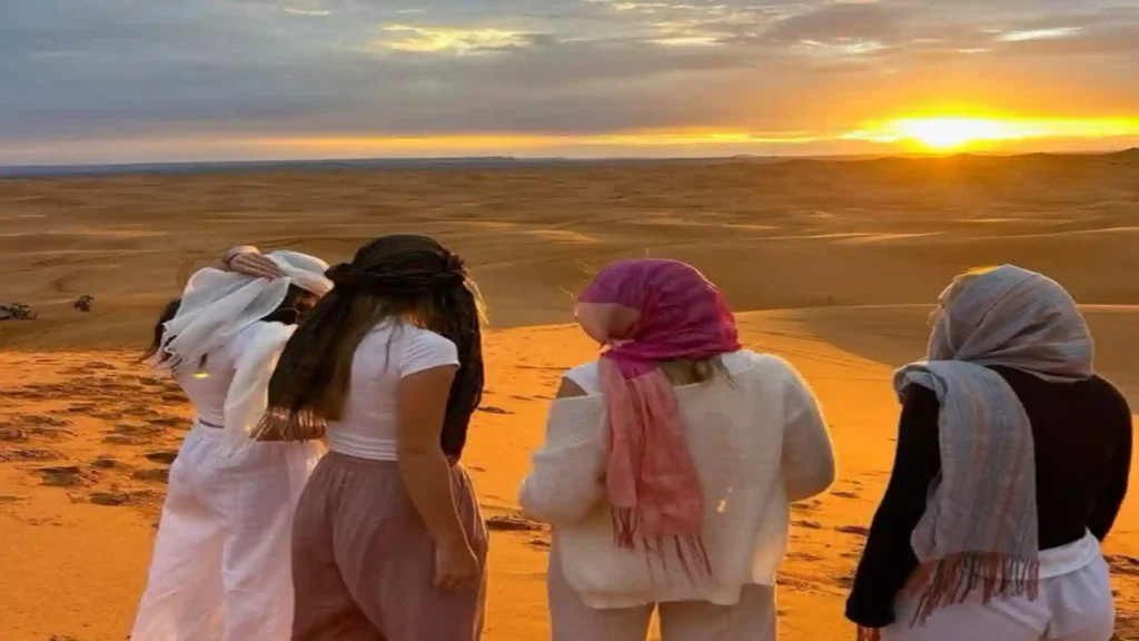 Four women viewed from behind sitting on the crest of a sand dune watching the bright yellow sun set over the Merzouga Morocco.