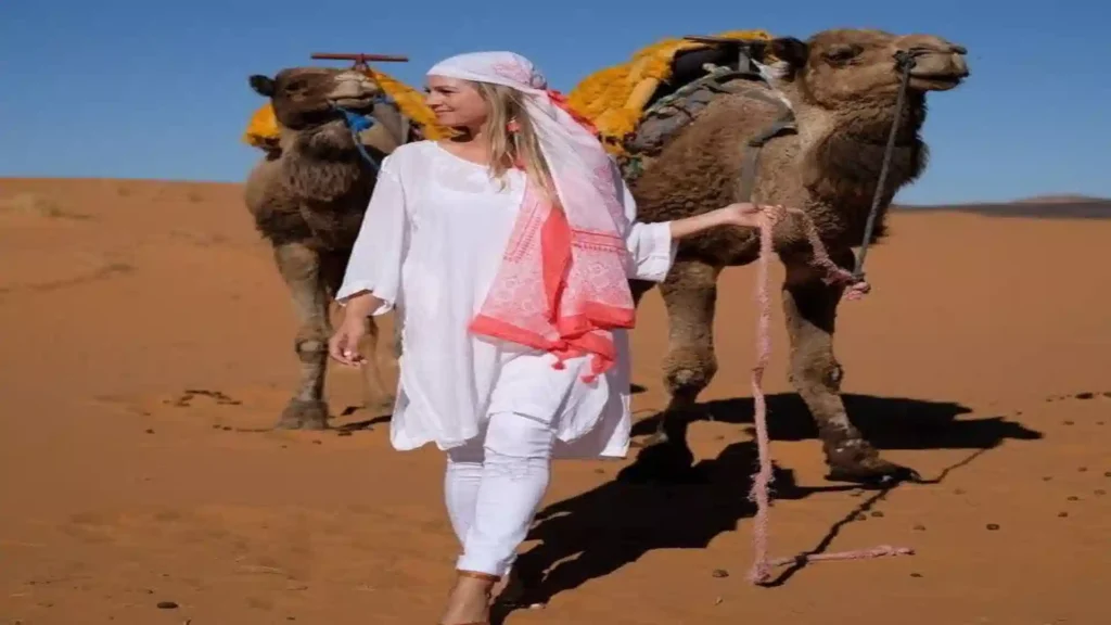 woman dressed in white with a headscarf leading two camels across the golden Erg Chebbi sand dunes in Merzouga Morocco.
