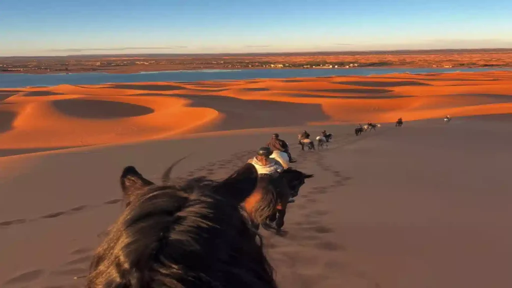 Point of view from a horse rider's perspective looking over a line of riders descending sand dunes at golden hour toward Dayet Srji lake. horse riding in Morocco